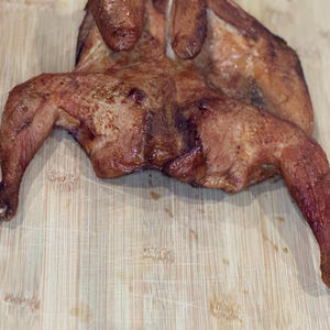 Könnte beinhalten: Ein ganzes gebratenes Hähnchen, goldbraun mit knuspriger Haut, wird auf einem hellen Holzschneidebrett präsentiert. Das Hähnchen ist perfekt gegart, mit sichtbaren Grillspuren und einem saftigen Aussehen, bereit für ein köstliches Essen.