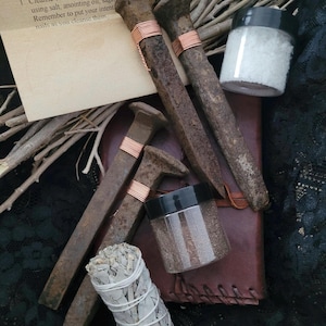May include: A set of rusty railroad spikes wrapped in copper wire, along with a small jar of white salt, a small jar of brown powder, and a bundle of white sage. The text on the card reads: "Follow these steps to help protect and bless your home using iron railroad spikes: 1. Cleanse each of the spikes. This can be using salt, anointing oil, sage. Remember to put your intent into the nails as you cleanse them."