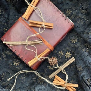 Könnte beinhalten: Ein handgefertigtes, dunkelrotes Lederjournal mit einer genähten Wirbelsäule, verziert mit Zimtstangen und Bindfaden. Das Journal ist mit einem helleren braunen Lederstreifen versehen. Der Hintergrund ist ein schwarzer Spitzenstoff.