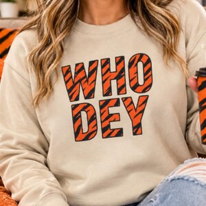 Op de afbeelding: Een beige sweater met ronde hals en de tekst WHO DEY in een vetgedrukt lettertype. De letters zijn gevuld met een oranje en zwart tijgerstreepmotief. De sweater heeft lange mouwen en een geribbelde halslijn, boorden en zoom.