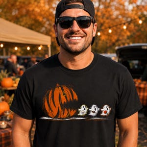 Op de afbeelding: Een zwart t-shirt met korte mouwen en een grafische print van een grote oranje uitgesneden pompoen die lijkt op een videogamepersonage en drie kleine witte geesten achtervolgt, elk met een sportgerelateerd voorwerp.