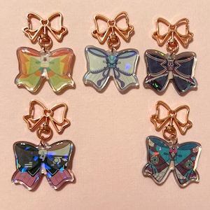 May include: Five iridescent butterfly-shaped keychains with gold-colored bow accents. Each keychain features a different butterfly design with various colors and patterns.