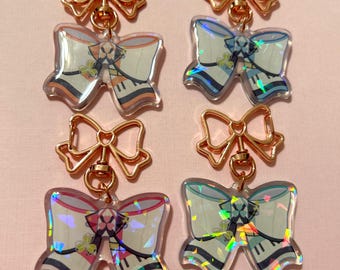 May include: Four iridescent holographic bow-shaped keychains with gold metal bow accents. Each keychain features a different design with a piano keyboard and floral accents.