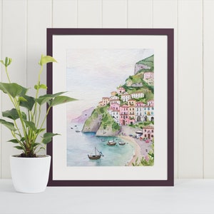 May include: Watercolor painting of a colorful Italian village nestled on a hillside overlooking a calm blue sea. The village is painted in shades of pink, yellow, and orange, with white accents. The sea is a light blue, and the sky is a soft, pale blue.