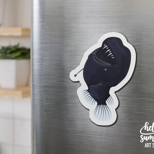 May include: A black and white anglerfish magnet with a large mouth and sharp teeth. The magnet is attached to a silver refrigerator.