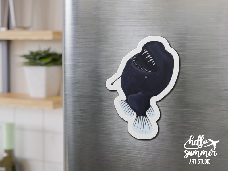 May include: A black and white anglerfish magnet with a large mouth and sharp teeth. The magnet is attached to a silver refrigerator.