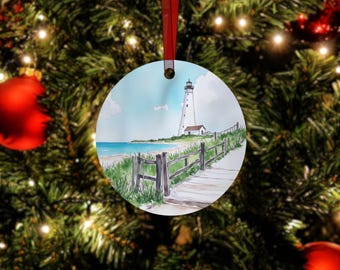 May include: A round white Christmas ornament with a watercolor painting of a white lighthouse on a beach with a wooden walkway leading to it. The lighthouse is on a grassy hill with a wooden fence.