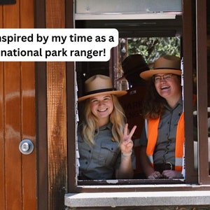 May include: Two women wearing ranger hats and uniforms smile and look out a window. The woman on the left is giving a peace sign. The text "inspired by my time as a national park ranger!" is written on a white background.