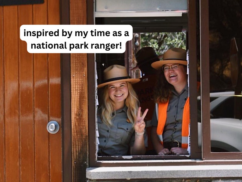 May include: Two women wearing ranger hats and uniforms smile and look out a window. The woman on the left is giving a peace sign. The text "inspired by my time as a national park ranger!" is written on a white background.