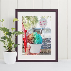 May include: A framed picture of a vibrant rainbow-coloured snow cone in a white paper cup. The image includes a sign that reads "Frost Bites". The snow cone is a mix of red, orange, yellow, green, and blue.