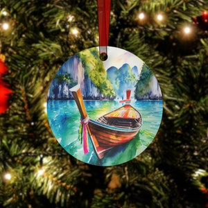 May include: A round Christmas bauble depicting a watercolour painting of a longtail boat in a tropical lagoon. The boat is brown with a red stripe and is surrounded by green cliffs and blue water.