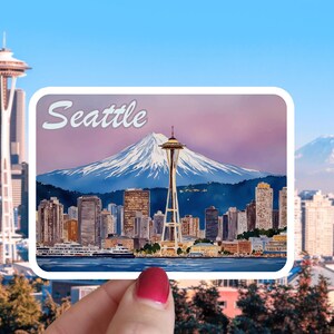 May include: A rectangular sticker featuring a watercolor painting of the Seattle skyline, Space Needle, and a snow-capped mountain. The word "Seattle" is written in white script across the top. The sticker is held up against a blurred background of the city.