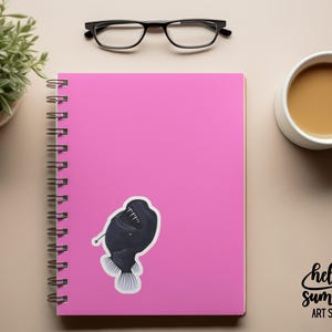 May include: A pink spiral bound notebook with a black and white anglerfish sticker on the front. The notebook is on a beige surface with a cup of coffee and a pair of glasses.