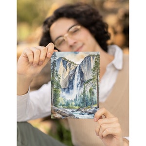 May include: A watercolor painting of Yosemite Falls in California. The painting depicts the waterfall cascading down a rocky cliff face, surrounded by lush green trees. The water is a light blue color, and the sky is a pale blue with hints of orange and yellow.