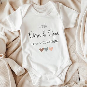 K&ouml;nnte beinhalten: Ein wei&szlig;er kurz&auml;rmeliger Babybody mit dem schwarzen Aufdruck "BEREIT Oma & Opa GENANNT ZU WERDEN?" und drei kleinen Herzen in gedeckten Braun-, Grau- und Pfirsicht&ouml;nen, die mittig unter dem Text auf der Vorderseite platziert sind.