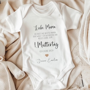Könnte beinhalten: Weißer Baby-Body mit kurzen Ärmeln. Der Body trägt den schwarzen Schriftzug "Liebe Mama" und "1. Muttertag". Der Text wird von einem kleinen Herz begleitet. Der Body liegt auf einer beigen Oberfläche.