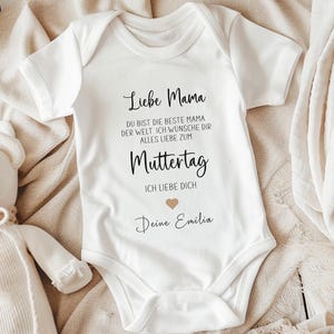 Pode incluir: Um body de bebé branco de manga curta com texto impresso em preto que diz: Liebe Mama, DU BIST DIE BESTE MAMA DER WELT, ICH WÜNSCHE DIR ALLES LIEBE ZUM Muttertag, ICH LIEBE DICH, seguido de um pequeno coração bege e o nome Deine Emilia.