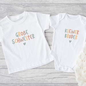 May include: A white short-sleeved t-shirt and a white short-sleeved baby bodysuit. The t-shirt features the text "GROBE SCHWESTER" in multicolored letters with a small heart. The bodysuit features the text "KLEINER BRUDER" in multicolored letters with a small heart.