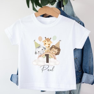 May include: A white short-sleeved t-shirt featuring a graphic of an elephant, giraffe, and lion wearing party hats. The animals are positioned above a rainbow and a large number one, with the name Paul written in black script below the design.