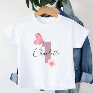 Può includere: Una t-shirt bianca a maniche corte con una grande grafica del numero uno in rosa tenue. Il design include una farfalla rosa posizionata sopra il numero e un fiore rosa chiaro sotto. Il nome Charlotte è stampato in corsivo nero.