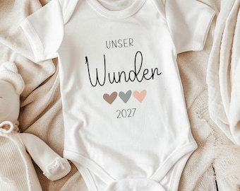 May include: A white short-sleeved baby bodysuit featuring the black text "UNSER Wunder" above three small hearts in brown, grey, and peach, with the number "2027" printed in black below the hearts.