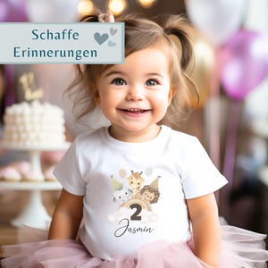 May include: A white t-shirt featuring a graphic of a cartoon elephant, giraffe, and lion wearing party hats above a cloud with the number 2 and the name Jasmin printed in black script. The text Schaffe Erinnerungen appears in a blue box.