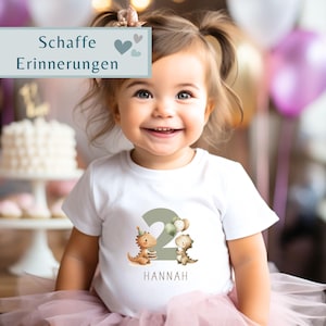 May include: A smiling toddler wearing a white t-shirt with a large number 2 and dinosaur graphics. The shirt also has the name "HANNAH" printed below the number. The child is wearing a pink tutu and has two pigtails. The text "Schaffe Erinnerungen" is in the background.