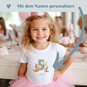 May include: A young girl wearing a white t-shirt with a dinosaur graphic and the number 5. The shirt also has the name "LILLY" printed on it. She is wearing a pink tutu and a pink crown. The text at the top says "Mit dem Namen personalisiert".