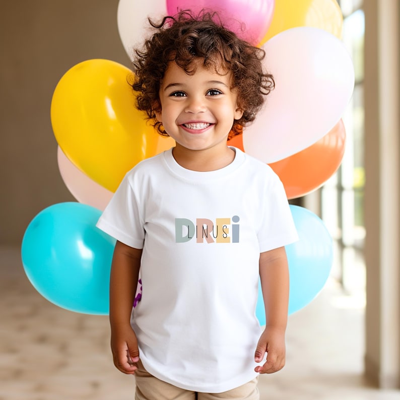 May include: A white short-sleeved t-shirt featuring the text DREI LINUS in multicolored pastel block letters across the chest. The shirt is worn by a child with curly brown hair, positioned in front of a cluster of colorful balloons.