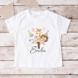 May include: A white short-sleeved t-shirt featuring a graphic of an elephant, giraffe, and lion wearing party hats. The animals are positioned above a rainbow and a cloud with the number 1 and the name Emilia printed in black script below.