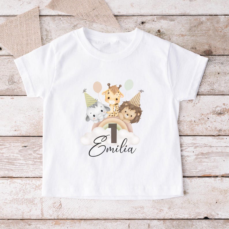 May include: A white short-sleeved t-shirt featuring a graphic of an elephant, giraffe, and lion wearing party hats. The animals are positioned above a rainbow and a cloud with the number 1 and the name Emilia printed in black script below.