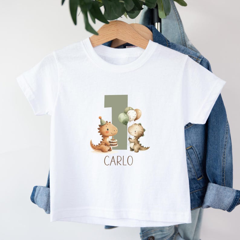 May include: White toddler t-shirt featuring a birthday design. The design includes two cartoon dinosaurs, the number 1, balloons, and the name "CARLO" in a simple font. The dinosaurs are holding a cake and balloons.