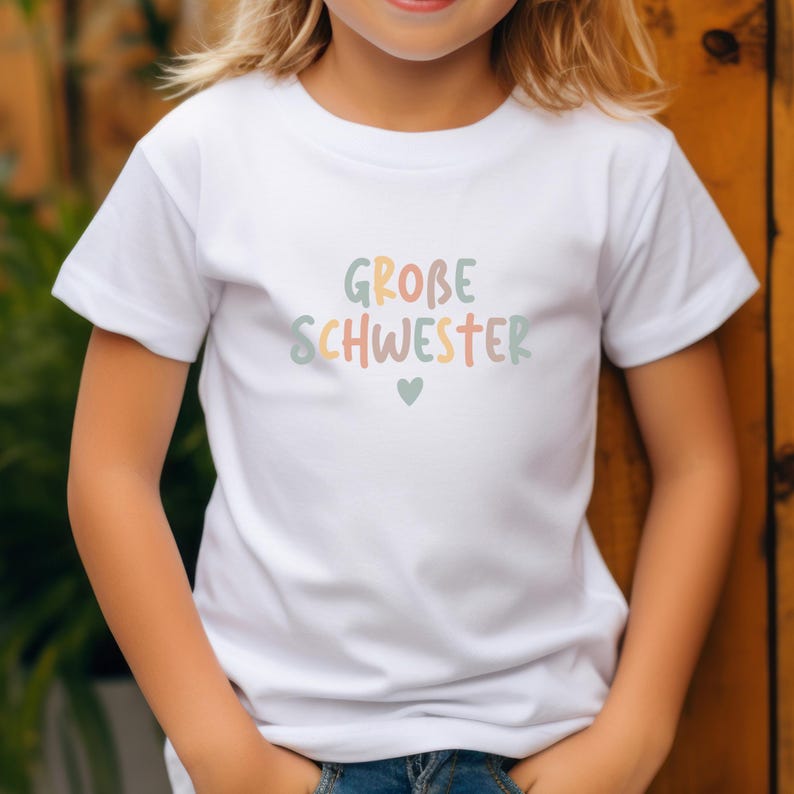 May include: A white short-sleeved t-shirt featuring the text GROBE SCHWESTER in multi-colored pastel letters across the chest, accompanied by a small heart icon centered below the text. The shirt has a crew neckline and a plain white fabric finish.