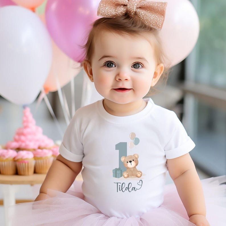 Peut inclure: Un body blanc &agrave; manches courtes orn&eacute; d'un motif repr&eacute;sentant le chiffre un bleu clair, un ours en peluche marron, deux petits ballons et un cadeau. Le pr&eacute;nom Tilda est inscrit en &eacute;criture cursive sous le motif, avec un petit c&oelig;ur dessin&eacute;.