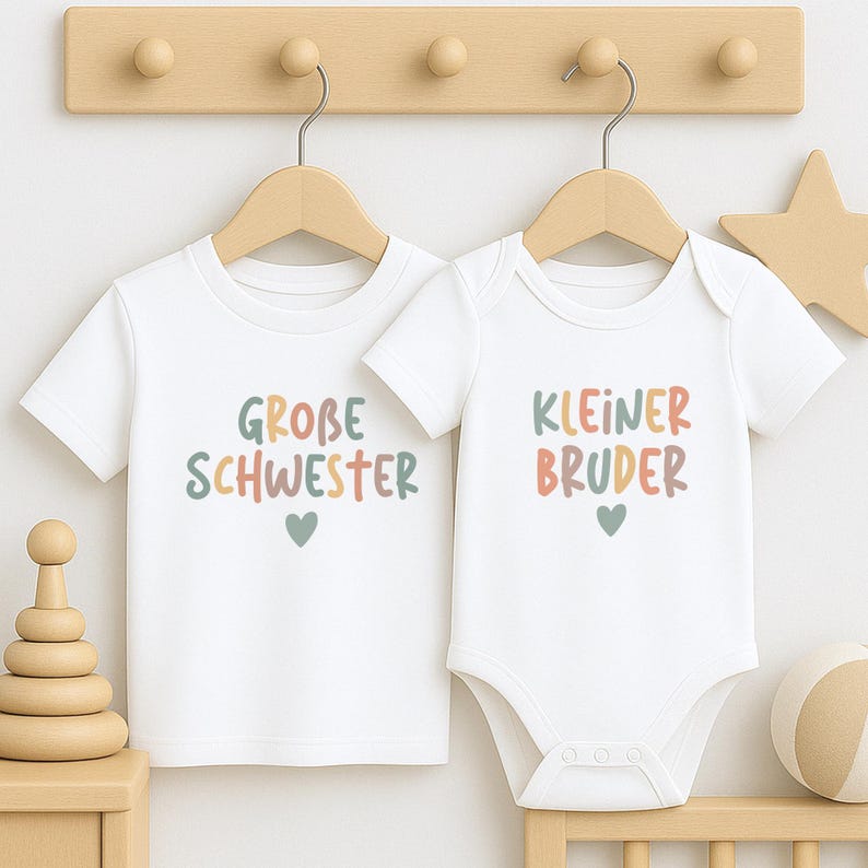 May include: A white short-sleeved t-shirt and a white short-sleeved bodysuit. The t-shirt features the text "GROBE SCHWESTER" in multicolored letters with a small heart below. The bodysuit features the text "KLEINER BRUDER" in multicolored letters with a small heart below.