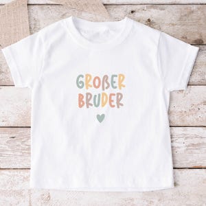 May include: A white short-sleeved t-shirt featuring the text GROBER BRUDER in a multi-colored, rounded sans-serif font. The letters are colored in muted shades of sage green, dusty orange, and tan. A small sage green heart is centered below the text.