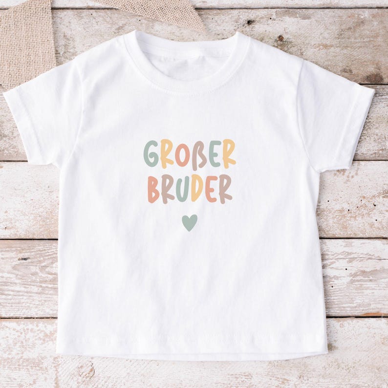 May include: A white short-sleeved t-shirt featuring the text GROBER BRUDER in a multi-colored, rounded sans-serif font. The letters are colored in muted shades of sage green, dusty orange, and tan. A small sage green heart is centered below the text.