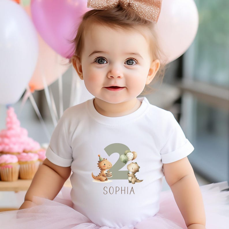 May include: A white t-shirt with a birthday theme. The shirt features a large number "2" in green, with two cartoon dinosaurs and the name "Sophia" printed below. The child is wearing a pink tutu and has a gold bow in their hair.