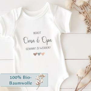 K&ouml;nnte beinhalten: Ein wei&szlig;er kurz&auml;rmeliger Babybody mit dem schwarzen Aufdruck BEREIT Oma & Opa GENANNT ZU WERDEN? und drei kleinen Herzen in gedeckten Braun-, Blau- und Rosat&ouml;nen, die mittig auf der Brust des Kleidungsst&uuml;cks platziert sind.