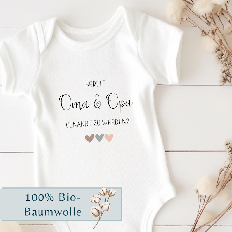 K&ouml;nnte beinhalten: Ein wei&szlig;er kurz&auml;rmeliger Babybody mit dem schwarzen Aufdruck BEREIT Oma & Opa GENANNT ZU WERDEN? und drei kleinen Herzen in gedeckten Braun-, Blau- und Rosat&ouml;nen, die mittig auf der Brust des Kleidungsst&uuml;cks platziert sind.