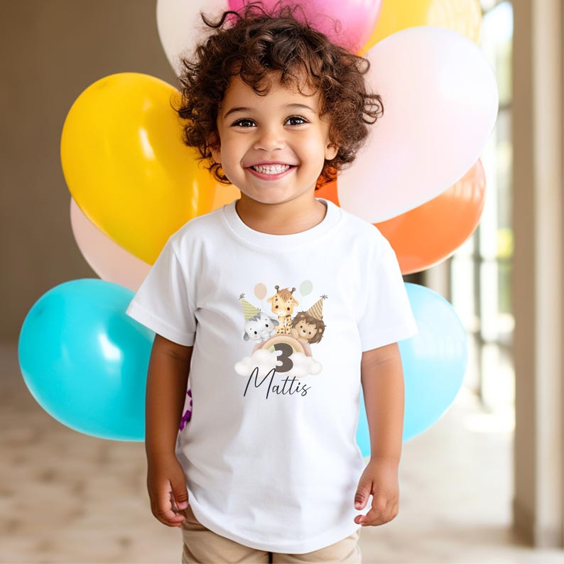 May include: A white short-sleeved t-shirt featuring a graphic of a cartoon elephant, giraffe, and lion wearing party hats above a cloud with the number 3 and the name Mattis printed in black script. The shirt is worn by a child with curly hair.