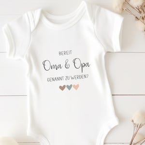K&ouml;nnte beinhalten: Ein wei&szlig;er kurz&auml;rmeliger Babybody mit dem schwarzen Aufdruck BEREIT Oma & Opa GENANNT ZU WERDEN? und drei kleinen Herzen in gedecktem Braun, Blaugrau und Hellrosa, die mittig auf der Vorderseite des Kleidungsst&uuml;cks platziert sind.
