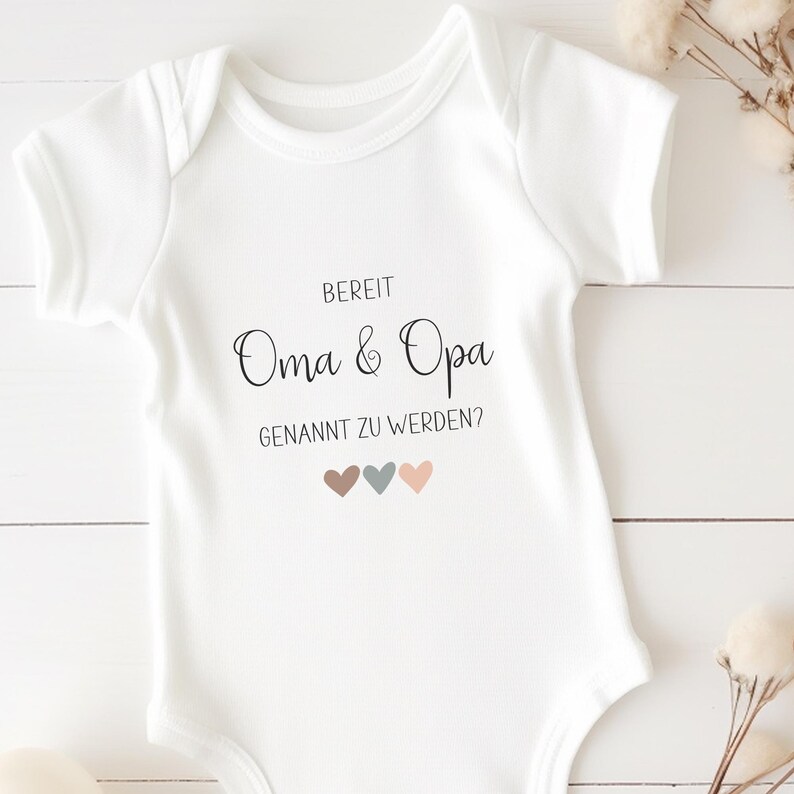 K&ouml;nnte beinhalten: Ein wei&szlig;er kurz&auml;rmeliger Babybody mit dem schwarzen Aufdruck BEREIT Oma & Opa GENANNT ZU WERDEN? und drei kleinen Herzen in gedecktem Braun, Blaugrau und Hellrosa, die mittig auf der Vorderseite des Kleidungsst&uuml;cks platziert sind.