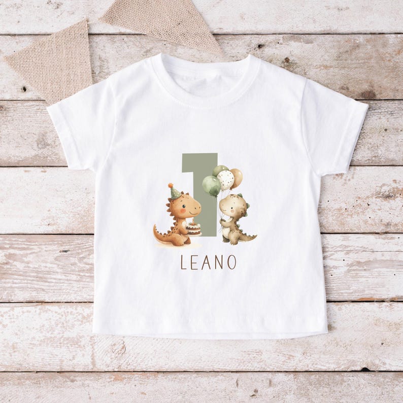 May include: White t-shirt featuring a birthday design. The design includes the number 1, two cartoon dinosaurs, balloons, and the name "LEANO" in a simple font. The dinosaurs are in shades of brown and green.