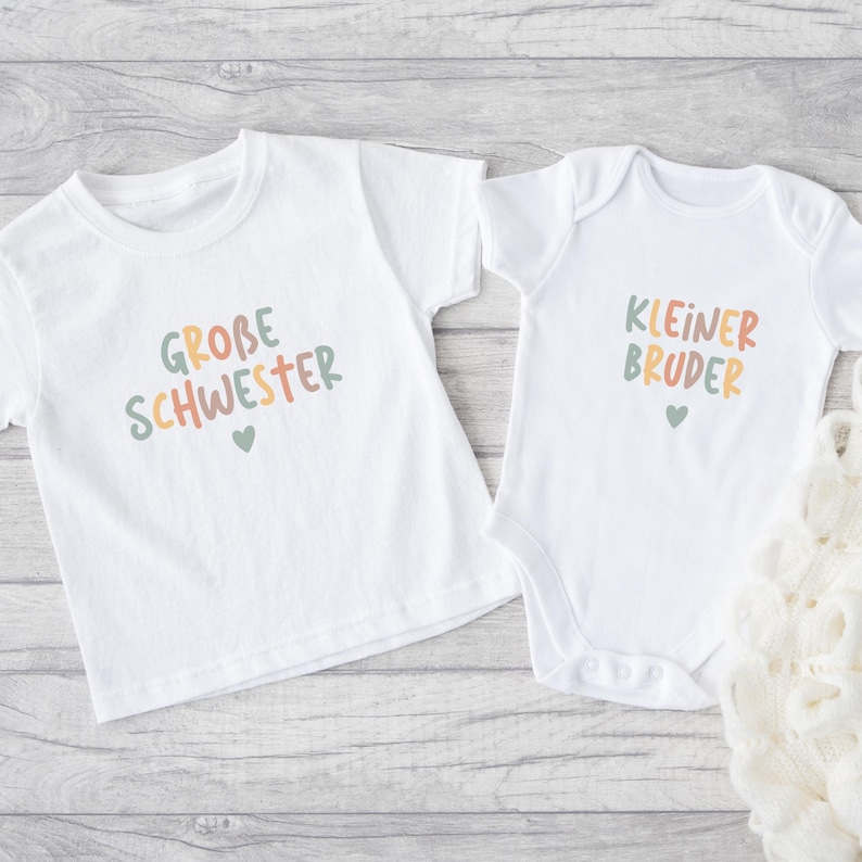 May include: A white short-sleeved t-shirt and a white short-sleeved baby bodysuit. The t-shirt features the text "GROBE SCHWESTER" in multicolored letters with a small heart. The bodysuit features the text "KLEINER BRUDER" in multicolored letters with a small heart.