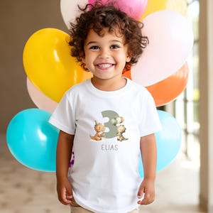 May include: A white t-shirt featuring a design for a third birthday. The design includes the number 3 in a muted green color, along with two cartoon dinosaurs and the name "ELIAS" in a simple font. The background includes colorful balloons.