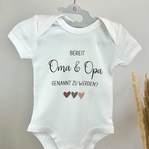 K&ouml;nnte beinhalten: Ein wei&szlig;er kurz&auml;rmeliger Babybody mit dem schwarzen Aufdruck "BEREIT Oma & Opa GENANNT ZU WERDEN?" und drei kleinen Herzen in Braun, Dunkelgrau und Hellrosa unter dem Text, der an einem grauen Kleiderb&uuml;gel h&auml;ngt.