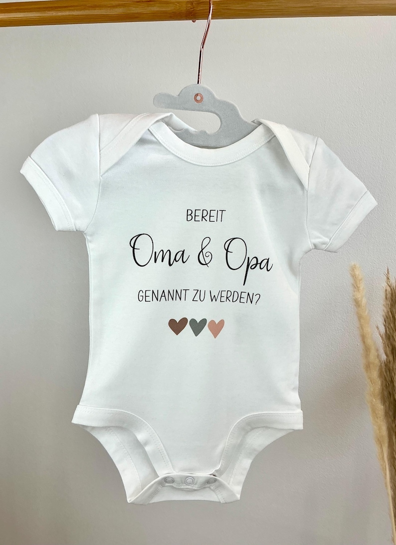 K&ouml;nnte beinhalten: Ein wei&szlig;er kurz&auml;rmeliger Babybody mit dem schwarzen Aufdruck "BEREIT Oma & Opa GENANNT ZU WERDEN?" und drei kleinen Herzen in Braun, Dunkelgrau und Hellrosa unter dem Text, der an einem grauen Kleiderb&uuml;gel h&auml;ngt.