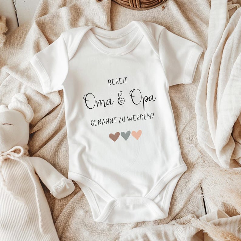 K&ouml;nnte beinhalten: Ein wei&szlig;er kurz&auml;rmeliger Babybody mit dem schwarzen Aufdruck "BEREIT Oma & Opa GENANNT ZU WERDEN?" und drei kleinen Herzen in gedeckten Braun-, Grau- und Pfirsicht&ouml;nen, die mittig unter dem Text auf der Vorderseite platziert sind.