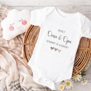 K&ouml;nnte beinhalten: Ein wei&szlig;er kurz&auml;rmeliger Babybody mit dem schwarzen Aufdruck "BEREIT Oma & Opa GENANNT ZU WERDEN?" und drei kleinen Herzen in den Farben Braun, Salbeigr&uuml;n und zartem Rosa, die mittig unter dem Text auf der Vorderseite platziert sind.